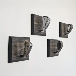 May include: Four golf club holders mounted on a white wall. Each holder is a square, stained wooden plaque with a black border, designed to display a golf club. The golf clubs are silver and black.