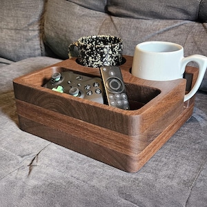 May include: A wooden sofa tray with a built-in cup holder and a compartment for a remote control and a game controller. The tray holds a white mug and a black and white speckled mug. The tray is made of dark brown wood.