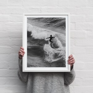 Könnte beinhalten: Schwarz-Weiß-Druck in einem Rahmen, der einen Surfer auf einer Welle zeigt. Der Surfer, in einem schwarzen Neoprenanzug, steht auf einem Surfbrett. Das Bild fängt die Kraft der Welle und das Können des Surfers ein. Der Rahmen ist weiß.