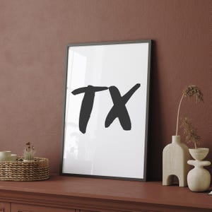 Może przedstawiać: Oprawiony wydruk z literami "TX" w odważnych, czarnych pociągnięciach pędzla na białym tle. Grafika jest prezentowana w czarnej ramie i opiera się o ciemnoczerwoną ścianę. Ozdobne wazony i pleciony kosz znajdują się na pierwszym planie.