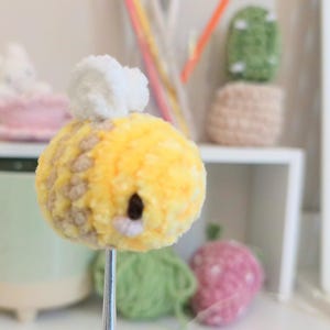 May include: A close-up of a yellow and brown crocheted bee with white wings. The bee is on a metal stick. Other crocheted items are in the background, including a pink bunny in a teacup, a green cactus, and a pink strawberry.