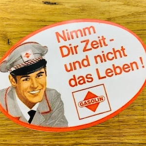 Könnte beinhalten: Ein weißer ovaler Aufkleber mit rotem Rand. Der Aufkleber zeigt einen lächelnden Mann in grauer Uniform und schwarzer Mütze. Der Text auf dem Aufkleber lautet "Nimm Dir Zeit - und nicht das Leben!" und "GASOLIN" in einer roten Raute.