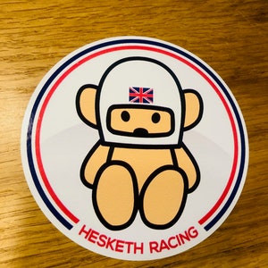 Pode incluir: Um adesivo circular branco com um urso de desenho animado usando um capacete branco com uma bandeira da Union Jack. O urso está sentado com as pernas cruzadas. O adesivo tem uma borda vermelha e azul e o texto "Hesketh Racing" na parte inferior.