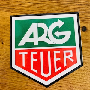 May include: A white sticker with a black border in the shape of a shield. The top half of the shield is green with the white text "ARG" and a white arrow pointing to the right. The bottom half of the shield is red with the white text "TEUER".