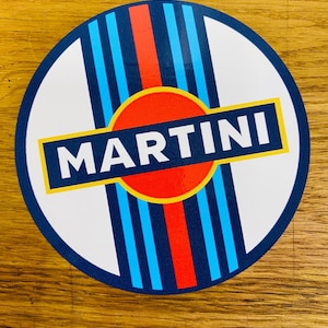 Puede incluir: Adhesivo redondo con el logotipo de Martini. Presenta un fondo blanco, rayas azules y rojas, y la palabra "MARTINI" en un rectángulo azul con borde amarillo. El adhesivo tiene un borde azul oscuro.