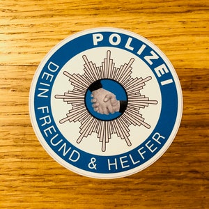 Puede incluir: Una pegatina redonda azul y blanca con la palabra "POLIZEI" en un círculo alrededor de una estrella blanca con un apretón de manos en el centro. El texto debajo de la estrella dice "DEIN FREUND & HELFER".