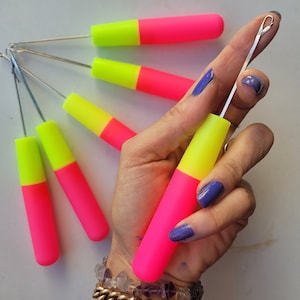 Peut inclure: Un ensemble de six aiguilles à tricoter de couleur rose vif et jaune fluo avec un crochet métallique à l'extrémité. Les aiguilles sont tenues dans une main avec un vernis à ongles bleu.