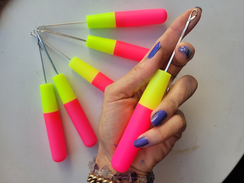 Peut inclure: Un ensemble de six aiguilles &agrave; tricoter de couleur rose vif et jaune fluo avec un crochet m&eacute;tallique &agrave; l'extr&eacute;mit&eacute;. Les aiguilles sont tenues dans une main avec un vernis &agrave; ongles bleu.