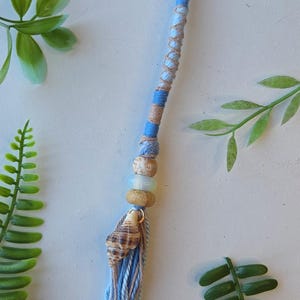 May include: A handmade tassel featuring a seashell, wooden beads, and a blue and tan wrapped cord. The tassel has a blue and tan yarn fringe. Green leaves are in the background.