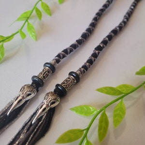 May include: Two black and silver braided hair ties with silver skull beads. The hair ties are on a white background with green leaves.
