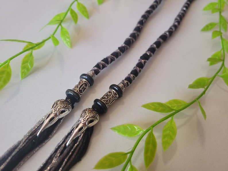 May include: Two black and silver braided hair ties with silver skull beads. The hair ties are on a white background with green leaves.