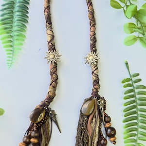 May include: Two hair wraps featuring braided tan and dark brown fabric strips, accented with metallic gold sun charms. The ends are decorated with dark brown wooden beads, smooth brown teardrop stones, and small metallic bird skull charms.