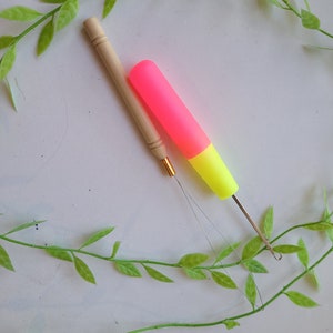 Peut inclure: Deux aiguilles &agrave; laine, l'une avec un manche en bois et l'autre avec un manche en plastique rose et jaune, sont pos&eacute;es sur une surface blanche avec des vignes vertes.