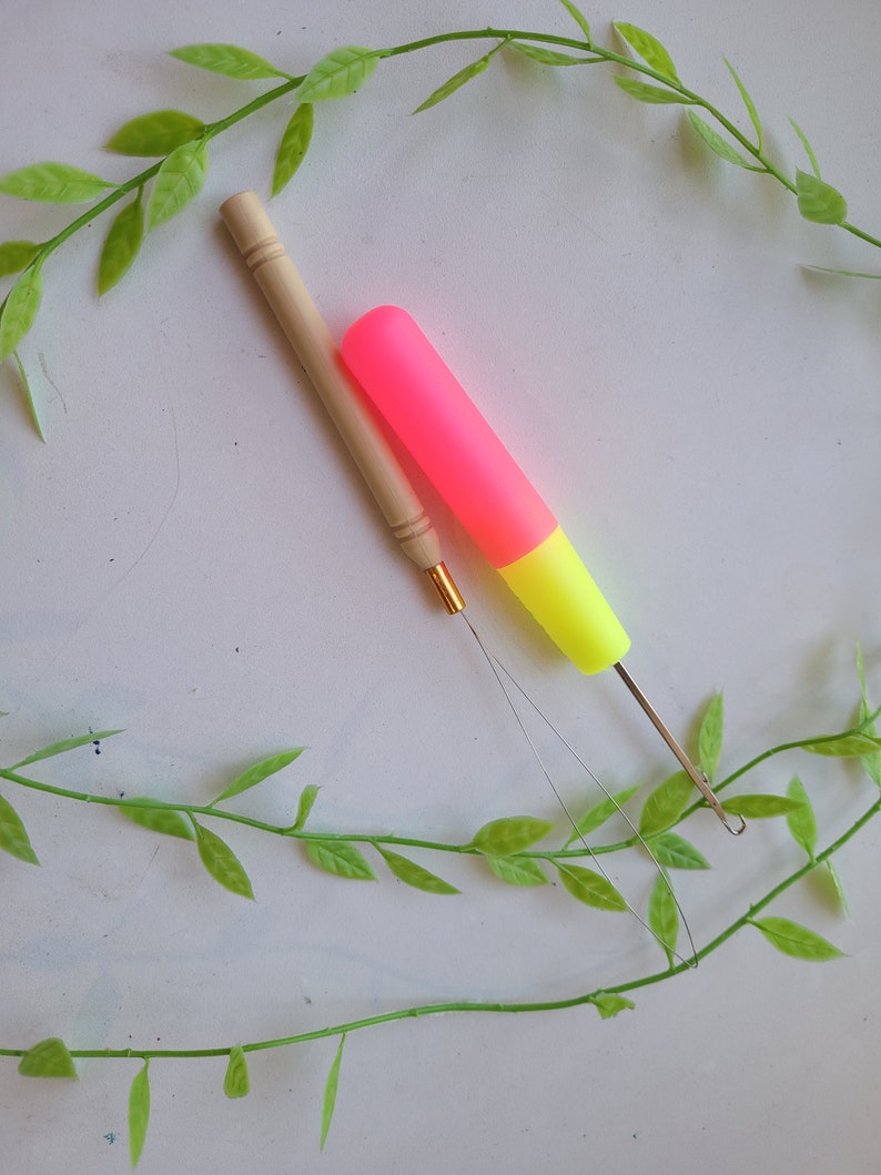 Peut inclure: Deux aiguilles &agrave; laine, l'une avec un manche en bois et l'autre avec un manche en plastique rose et jaune, sont pos&eacute;es sur une surface blanche avec des vignes vertes.