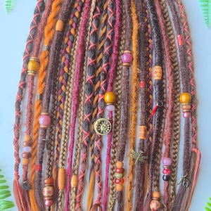 May include: A selection of vibrant dreadlocks in brown, orange, and pink, decorated with beads and charms. The dreads feature detailed braiding and orange thread wrapping. Charms include a sun, dragonfly, and heart. The overall style is bohemian.