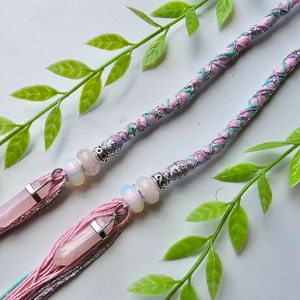 Puede incluir: Dos extensiones de cabello hechas a mano con detalles en rosa, plata y turquesa. Cada extensión presenta un colgante de cristal de cuarzo rosa, cuentas y borlas plateadas. Las extensiones están decoradas con tela trenzada y adornos plateados.