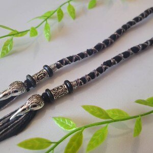 May include: A black and silver braided cord with two silver skull-shaped beads and tassels. The cord is lying on a white surface with green leaves.