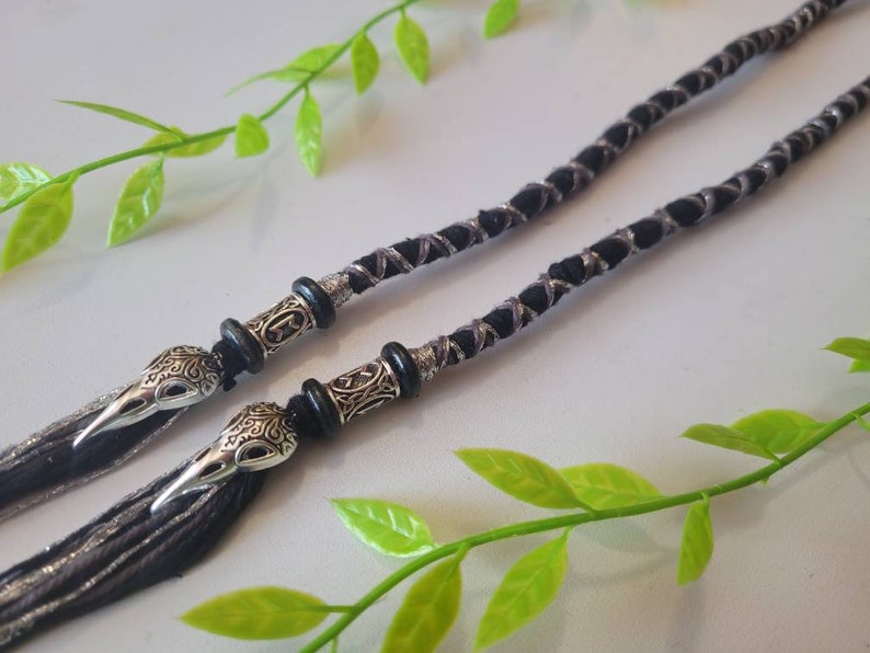 May include: A black and silver braided cord with two silver skull-shaped beads and tassels. The cord is lying on a white surface with green leaves.