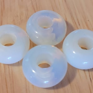 Puede incluir: Cuatro cuentas de opalo blanco en forma de rosquilla con un agujero en el centro. Las cuentas son lisas y tienen un ligero brillo iridiscente.