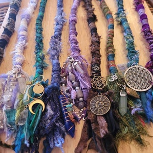 May include: A collection of eight colorful, handmade hair extensions with various charms and beads. The extensions are made of yarn and fabric and feature a variety of colors, including purple, blue, green, and brown. The charms include a tree of life, a sun, a moon, and a flower of life.