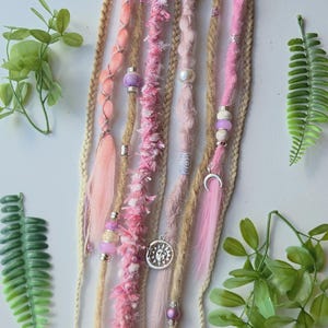 May include: A set of hair extensions featuring a mix of braided strands and loose fibres in shades of pink, peach, and beige. The strands are decorated with various beads, including purple and white spheres, and silver charms shaped like a sun, moon, and feather.
