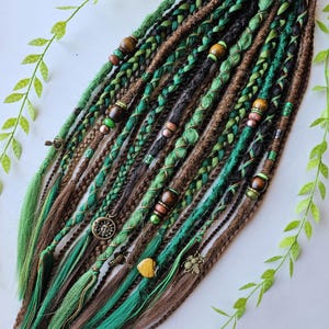 Op de afbeelding: Groene en bruine synthetische dreadlocks met kralen en bedels. De dreadlocks zijn bij elkaar gebonden met een zwart elastiek.