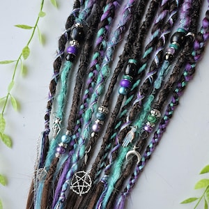 Puede incluir: Un conjunto de rastas sintéticas con detalles azules, morados y plateados. Las rastas están decoradas con cuentas, abalorios y colgantes, incluyendo un pentagrama, una luna creciente y una mano.