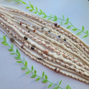 May include: A set of white synthetic dreadlocks with brown, red, and white beads. The dreadlocks are arranged in a row on a white surface.