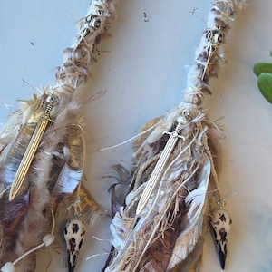 Peut inclure: Deux wraps de dreadlocks faits à la main, d'esthétique bohème. Chaque wrap est composé de tissus beiges, bruns et blancs, ornés de breloques épées argentées, de perles de crâne et d'accents de nœuds celtiques. Accessoires capillaires.