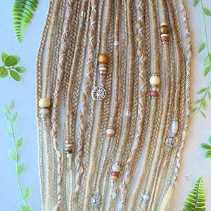 Könnte beinhalten: Ein Set aus blonden und braunen synthetischen Dreadlocks mit weißen und braunen Akzenten. Die Dreadlocks sind mit Perlen, Charms und Federn verziert.