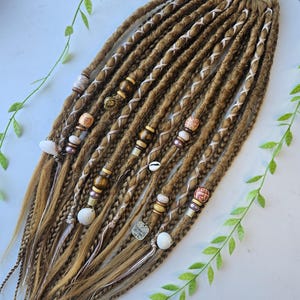 May include: A set of brown and blonde synthetic dreadlocks with white and brown string accents. The dreadlocks are decorated with beads, shells, and charms.  The dreadlocks are tied together with a brown elastic band.
