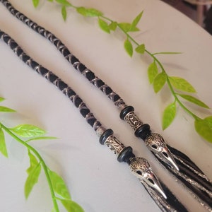 May include: Two black and silver braided cords with silver bird-shaped charms at the ends. The cords are hanging from a white surface.