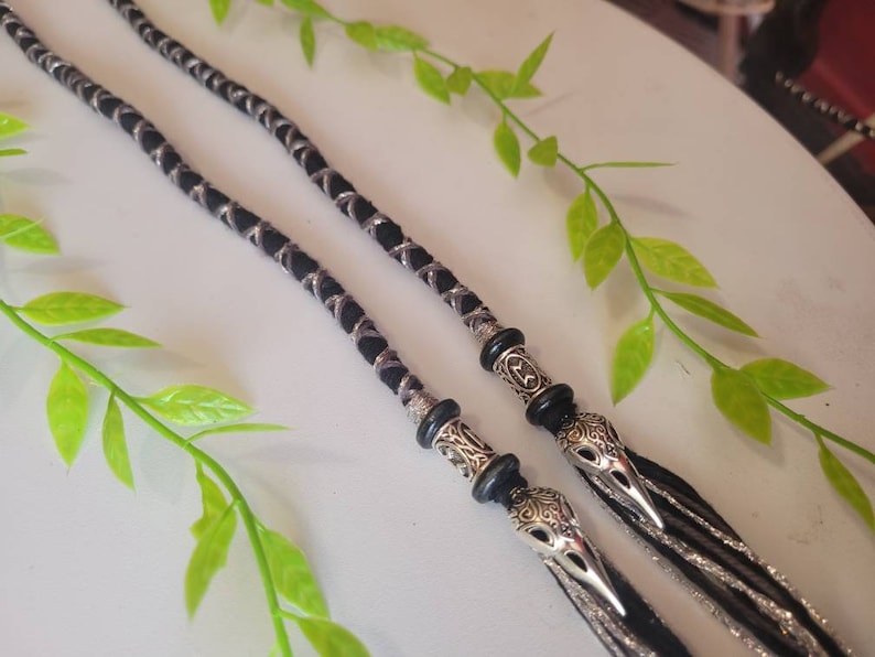 May include: Two black and silver braided cords with silver bird-shaped charms at the ends. The cords are hanging from a white surface.
