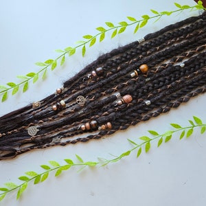 Op de afbeelding: Een set bruine synthetische dreadlocks met bruine en zilveren kralen en bedels. De dreadlocks zijn gerangschikt op een wit oppervlak met groene bladeren.