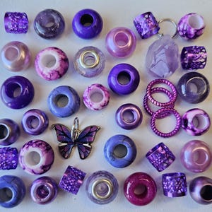 May include: A collection of purple and lavender beads, charms, and spacers. The beads are made of various materials, including glass, metal, and plastic. There is a butterfly charm and a purple crystal.