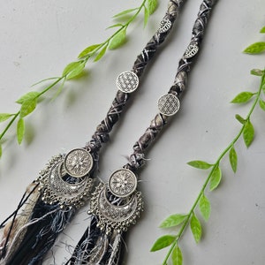 May include: Two black and white braided hair extensions with silver metal charms. The charms feature a flower design and a crescent moon shape. The extensions have tassels at the ends.