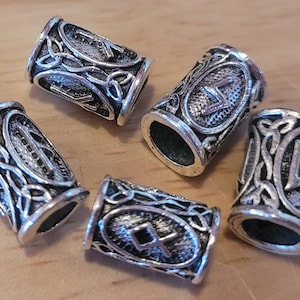 Puede incluir: Un conjunto de cuentas de metal plateado con intrincados diseños de nudos celtas y runas. Las cuentas cilíndricas presentan patrones y símbolos detallados, perfectos para la creación de joyas o proyectos de artesanía.