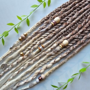 Peut inclure: Un ensemble de dreadlocks synthétiques brunes et blondes avec des perles et des breloques. Les dreadlocks sont disposées en rangée sur un fond bleu clair.