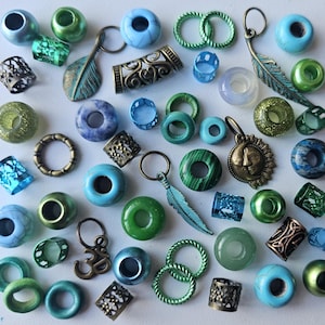 Op de afbeelding: Een assortiment van kleurrijke kralen en bedels in tinten blauw, groen en bruin. De kralen zijn gemaakt van verschillende materialen, waaronder glas, metaal en steen. Sommige kralen hebben ingewikkelde ontwerpen, terwijl andere eenvoudig zijn. De bedels omvatten een veer, een zon en een symbool.
