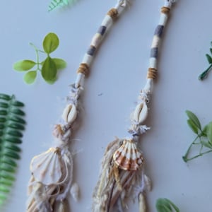 May include: Two hair wraps featuring white, grey, and tan fabric strips wrapped around a core. Each wrap is decorated with a white cowrie shell, a fan-shaped seashell, and a tassel made of frayed fabric strips and small dangling shell accents.