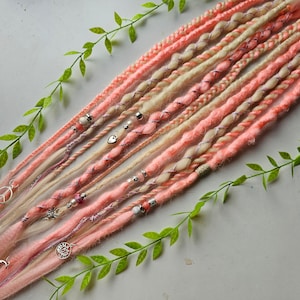 May include: A set of pink and blonde synthetic dreadlocks with silver accents and charms. The dreadlocks are arranged on a white surface with green leaves.