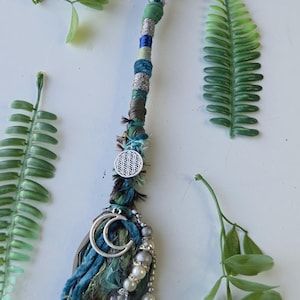 Puede incluir: Un accesorio para el cabello con un cordón largo trenzado en tonos azul, verde y plata. Está adornado con un dije plateado de la flor de la vida, un colgante de media luna, cuentas de perlas blancas y una punta de cristal verde oscuro.