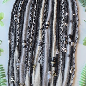 May include: A set of synthetic hair extensions featuring a mix of black, charcoal grey, and white dreadlocks and braids. The strands are adorned with various metallic charms, including pentagrams, crescent moons, and beads in silver and black tones.