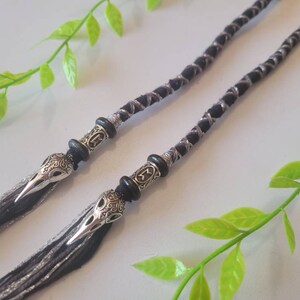 May include: Two black and silver braided cords with silver skull beads at the ends. The cords are lying on a white surface with green leaves.