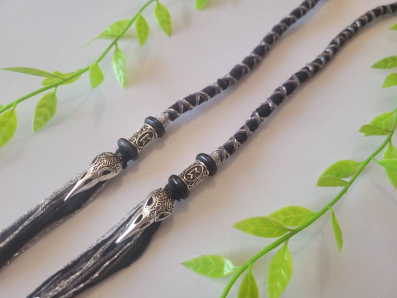 May include: Two black and silver braided cords with silver skull beads at the ends. The cords are lying on a white surface with green leaves.