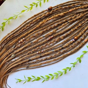 Puede incluir: Un conjunto de rastas de color marrón claro, adornadas con cuentas decorativas y envolturas de alambre. Las rastas están dispuestas sobre una superficie blanca, con ramitas verdes en los bordes. Las rastas tienen una mezcla de texturas y colores.