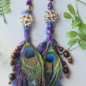 May include: Two hair extensions featuring wrapped purple, blue, and green fabric strands, accented with metallic sun-shaped charms containing clear crystals. Each extension includes a peacock feather, dark brown wooden beads, and frayed yarn.