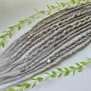Puede incluir: Una colección de rastas grises claras, adornadas con detalles plateados y cuentas decorativas. Algunas rastas presentan dijes plateados, incluyendo un árbol de la vida y diseños de hojas. Las rastas están dispuestas sobre una superficie blanca, con ramitas de hojas verdes.