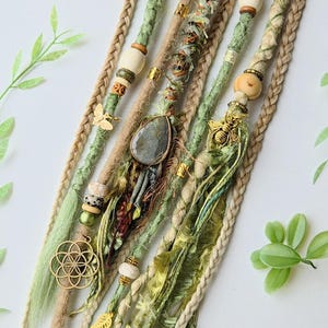 May include: A set of green and beige synthetic dreadlocks with various charms and beads. The charms include a gold flower of life, a gold bee, a gold leaf, and a grey stone. The beads are wooden and gold.