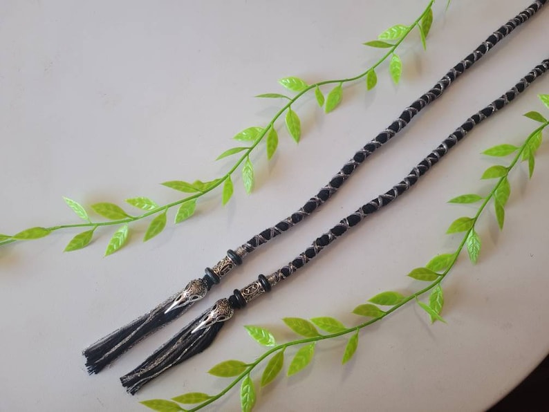 May include: Two black and silver braided cords with silver metal tassels. The cords have a decorative design and are tied together with a knot.